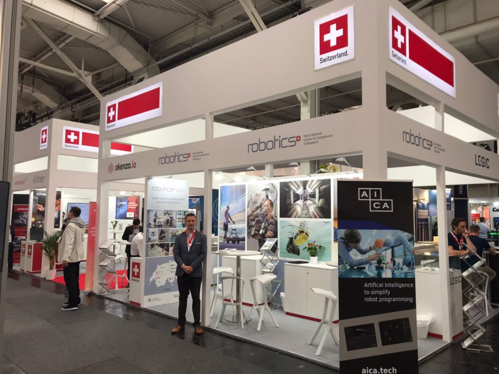 The image shows the NCCR Robotics booth at Hannover Messe and Michael smiling.