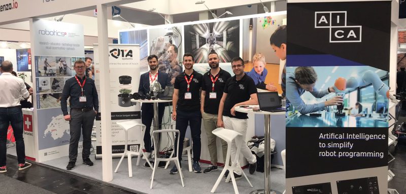 The image shows a booth of NCCR Robotics at Hannover Messe with banners from spin-offs AICA and BOTA SYSTEMS as well as 5 members of the team.